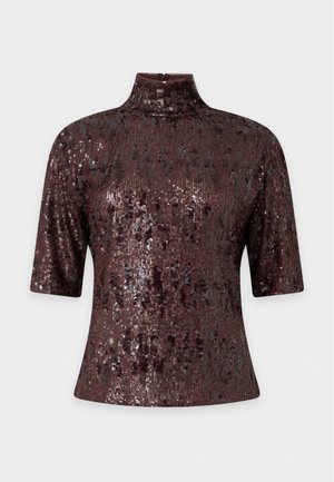 Une femme porte un haut sombre à sequins, une jupe bordeaux bordée de dentelle, des talons noirs et un sac porté à l’épaule, devant un fond uni.