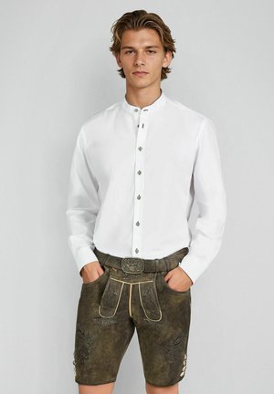 Junger Mann trägt traditionelle braune Lederhosen mit Stickerei und ein weißes Hemd mit Knöpfen, steht mit den Händen in den Taschen.