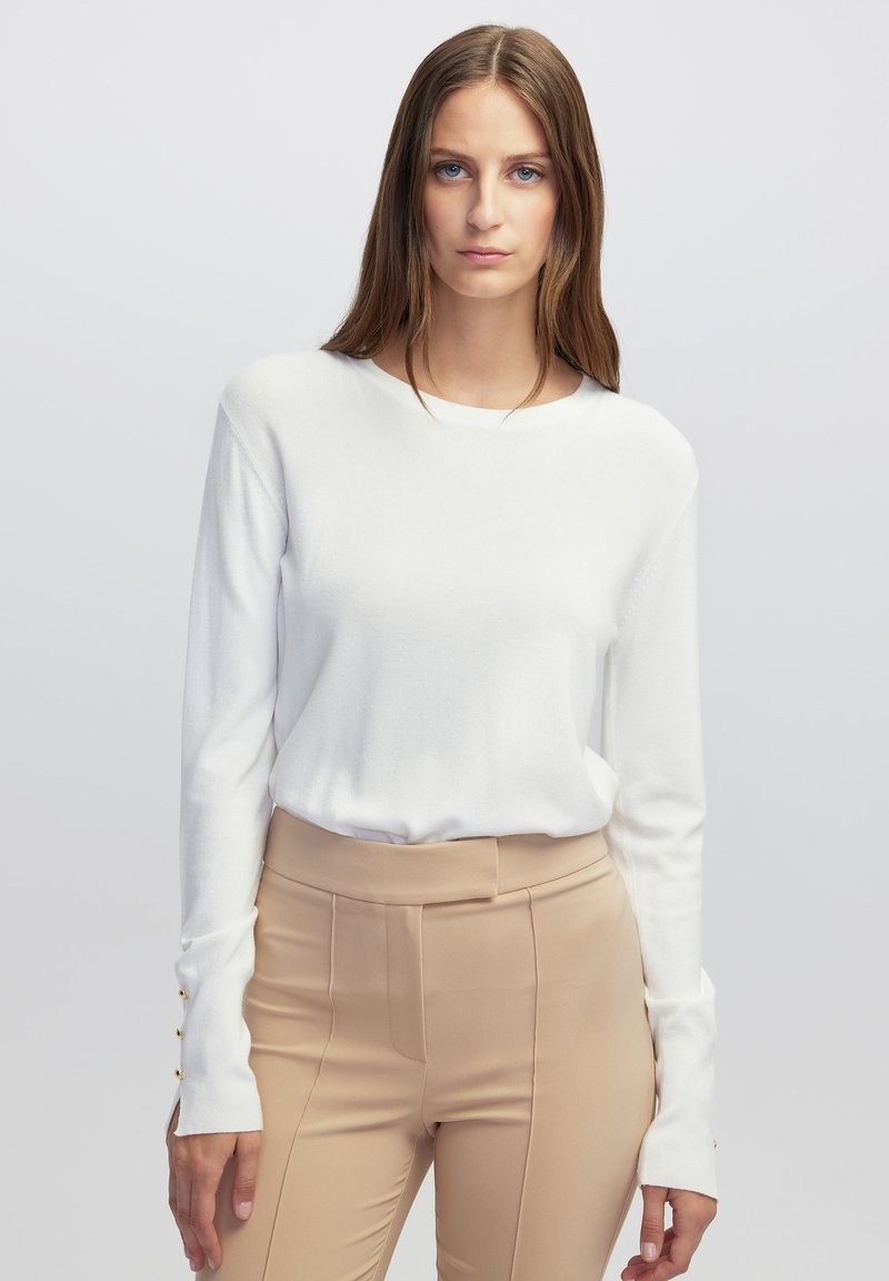 Haut en tricot blanc à manches longues avec un col rond et des accents de boutons sur les manches, associé à un pantalon haut de taille beige avec une texture lisse.
