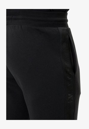 Pantalon de survêtement noir en tissu doux, doté d'une ceinture côtelée, de poches latérales et d'un motif logo ton sur ton le long du côté.