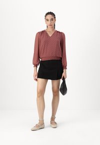 Blusa color ruggine con maniche a palloncino e orlo arricciato, abbinata a una mini gonna nera e scarpe piatte beige. Accessoriata con una borsa nera.