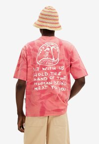 Růžové tričko s krátkým rukávem s efektom tie-dye, které má na zádech bílý grafický text a design. Spárované s tkaným kbelíkovým kloboukem s vícetrabnovým páskem.