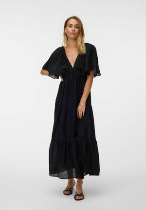 Femme portant une robe longue noire fluide avec un profond décolleté en V et des manches courtes larges, debout avec une main sur la taille et des sandales à talons noires.