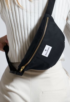 Sort canvas skuldertaske med en gylden lynlås, der har et logo-patch. Den justerbare strop er lavet af stof, og tasken har en buet form.