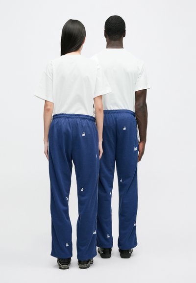 Marinblå joggingbyxor med vit kaninbrodering, elastisk midja och avslappnad passform; kombinerade med enfärgade vita t-shirts på två figurer sedda bakifrån.