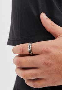 Sølvring med indgraveret tekst, bånddesign og indviklede detaljer, båret på en hånd mod en sort skjorte. Hånden viser naturlig tekstur.