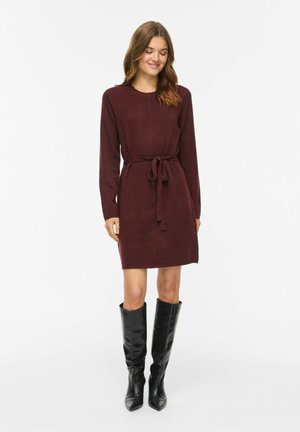 Femme portant une robe en maille bordeaux à manches longues avec une ceinture à la taille et des bottes noires hautes jusqu'aux genoux, debout devant un fond uni.
