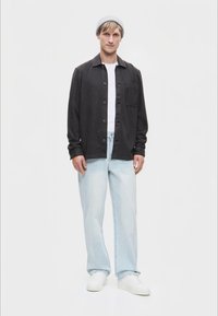 Σκούρο γκρι πουκάμισο πάνω από άσπρη μπλούζα, συνδυασμένο με ανοιχτό μπλε τζιν και άσπρα αθλητικά παπούτσια, συμπληρωμένο με γκρι σκουφάκι.