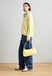 Gelbe Handtasche mit geschwungener Form und Obergriff, kombiniert mit einem flauschigen gelben Pullover, blauen weit geschnittenen Jeans und weißen Sneakers mit schwarzen Akzenten.