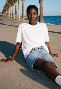 Young woman in white t-shirt and denim shorts sits on a promenade with palm trees and sea in the background.