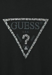 Must värvustatud kangas on kolmnurkne, kristallidega kaunistatud logo, kus on kirjad "GUESS" ja "U.S.A.", sealhulgas "WASHED JEANS" ning küsimärgi kujundus.