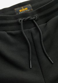 Un pantalon de jogging noir doté d'une ceinture côtelée avec cordon de serrage, d'embouts métalliques et d'une étiquette de produits durables cousue à l'intérieur.