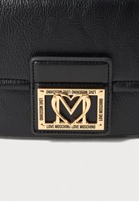 Melna teksturēta ādas soma ar zelta taisnstūrveida emblēmu, kurā ir sirds formas "M" un teksts "Love Moschino" uz visām pusēm.