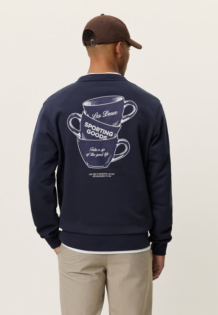 Φούτερ από φλις σε ναυτικό μπλε, με γραφικό σχέδιο στοιβαγμένων φλιτζανιών καφέ. Μακριά μανίκια, ριμπ μανσέτες και χαλαρή εφαρμογή.