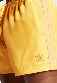 Sárga sportnadrág sima textúrával, rózsaszín oldalsávokkal és hímzett Adidas logóval az alján. Rugalmas derékpánt, kihúzható zsinórral.