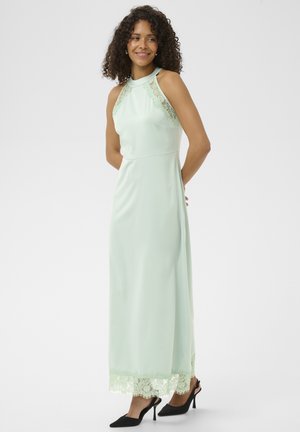 Kobieta uśmiechająca się, ubrana w bezrękawnikową, bladoniebieską suknię maxi z koronkowymi detalami oraz czarne szpilki, stojąca na jednolitym tle.