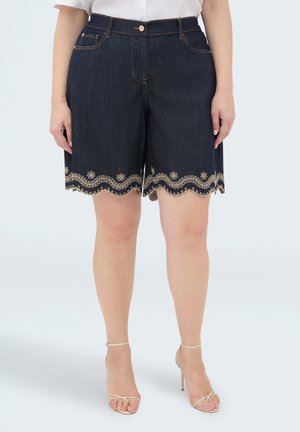 Ciemne szorty dżinsowe z beżowym kwiatowym haftem wzdłuż falowanych brzegów, noszone z beżowymi sandałami na wysokim obcasie i białą bluzką.