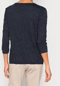 Granatowa bluzka z długim rękawem o pomarszczonej teksturze i szerokim dekoltem, zestawiona z jasnymi, miękkimi spodniami.