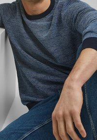Sweter wełniany w ciemnoniebieskim kolorze z teksturowanym wzorem, okrągłym dekoltem i ściągaczami. Noszony z niebieskimi dżinsami, z widoczną częścią ręki swobodnie spoczywającą.
