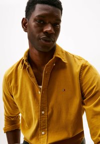 Uomo di colore che indossa una camicia a bottoni in velluto a coste color giallo senape con le maniche arrotolate e un'espressione seria, su uno sfondo bianco semplice.