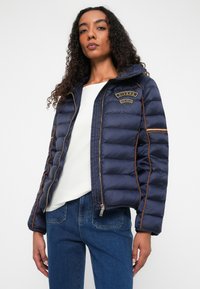 Marineblå puffjakke med gyldent branding, vandret syninger og orange piping. Har en lynlås lukning og hætte. Bæres over en hvid top.