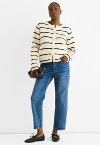 Cardigan a righe in crema e nero, con texture morbida, scollatura rotonda e chiusura a bottoni. Abbinato a jeans blu a gamba dritta e ballerine nere.