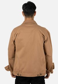 Chaqueta de lona marrón con cuello clásico, puños abotonados y dobladillo ajustable, que presenta una textura suave y un diseño de corte recto.