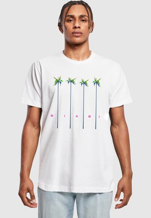 Mann iført hvit T-skjorte med fem tynne palmer og rosa bokstaver som staver "MIAMI" over brystet, sammen med lyseblå jeans.