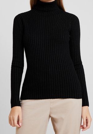 Personne portant un pull à col roulé côtelé noir à manches longues et un pantalon beige sur un fond uni.