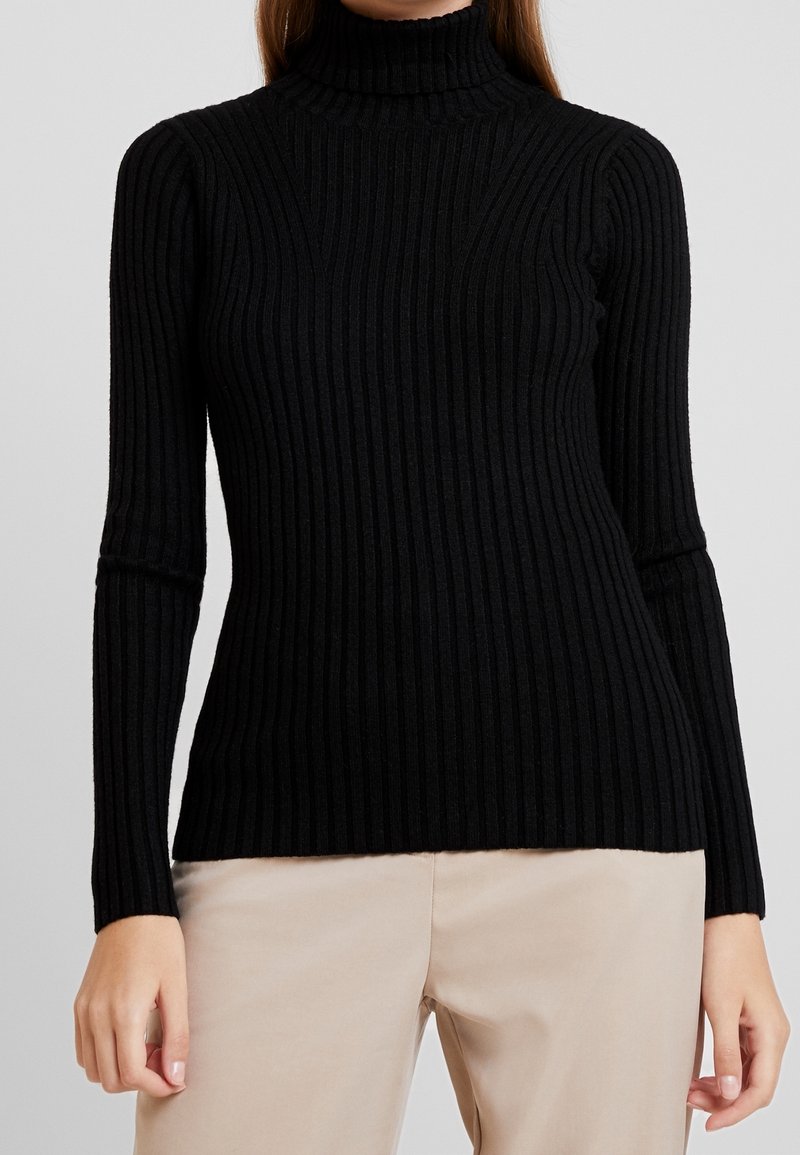 Personne portant un pull à col roulé côtelé noir à manches longues et un pantalon beige sur un fond uni.