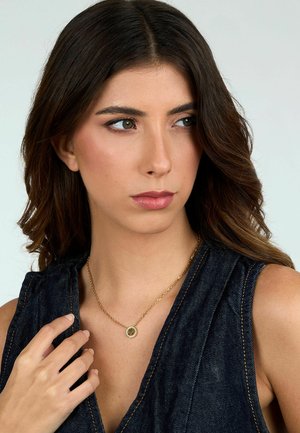 Collana d'oro con un pendente circolare, caratterizzato da dettagli intricati, indossata su una maglietta in denim scuro. Texture liscia e finitura lucida.