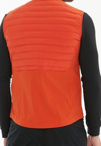 Vadderad orange väst med en slät textur och rundad nederkant, med quiltad sömnad på ryggen och svarta långärmade undertröjor under.