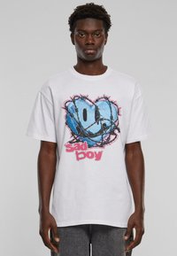 Hvid oversized t-shirt med et blåt smiley-ansigt grafik indkapslet i pigtråd og pink "sad boy" tekst nederst.
