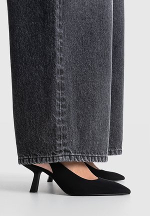 Schwarze Mules mit spitzer Kappe und Kitten-Heel, getragen zu weit geschnittenen dunkelgrauen Denim-Jeans, seitliche Ansicht auf Knöchelhöhe auf hellem Untergrund.
