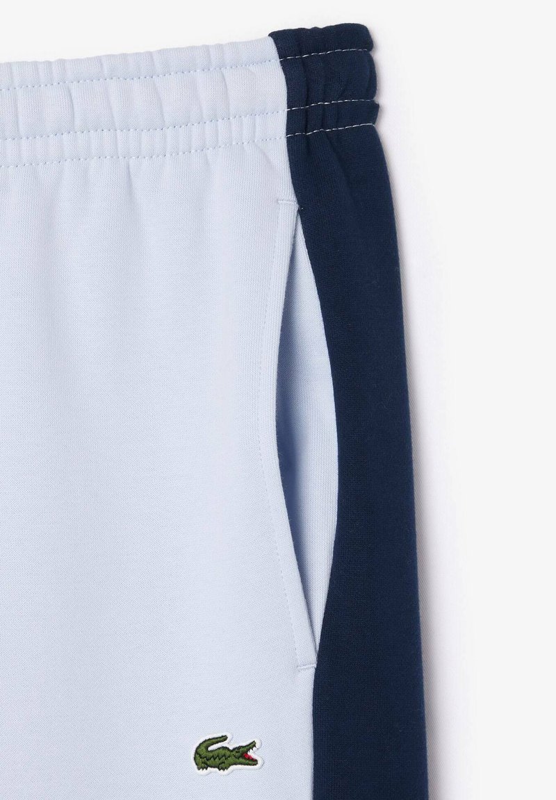 Pantalons de survêtement bleu clair avec un panneau latéral bleu foncé, taille élastique et logo de crocodile vert brodé près de la poche. Matériau en mélange de coton.