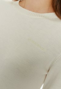 Maglione a maglia color crema con scollo a coste e logo "Guess Jeans" in rilievo sulla parte anteriore. Texture morbida, design aderente.