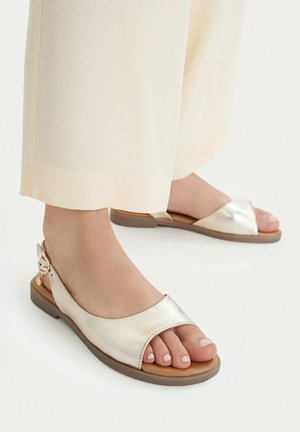 Füße, die flache, metallische, offene Slingback-Sandalen tragen, kombiniert mit cremefarbenen, weit geschnittenen Hosen vor weißem Hintergrund.