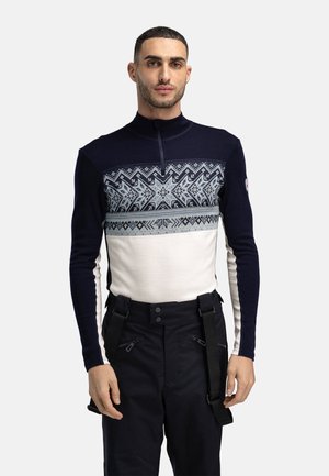 Mann iført en marineblå og hvit genser med glidelås og et nordisk mønster over brystet, samt svarte bukser med seler.