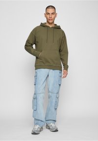 Oliiviroheline kapuutsiga dressipluus esitasku ja reguleeritavate nööridega, kombineeritud helesiniste küljetaskutega cargo-teksadega. Hallid spordijalatsid.