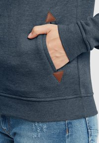 Mørkegrå hettegenser med teksturert overflate, utstyrt med kengurulomme og brun lærtriangel ved lomma.