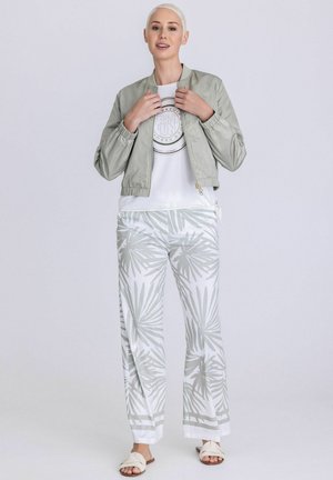 Frau mit kurzen blonden Haaren trägt eine hellgraue Bomberjacke, weißes T-Shirt mit Grafik, weiße Hose mit grauem Blattmuster und weiße Sandalen.