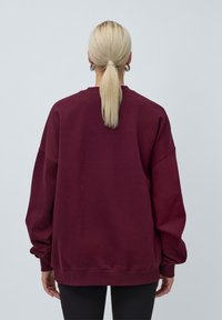 Burgunderfarget hettegenser med senkede skuldre og lange, løse ermer. Har en ribbestrikket rund hals og mansjetter, laget av myk teksturert stoff.