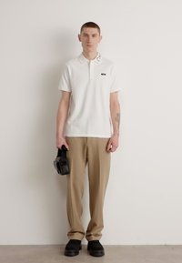 Witte polo met korte mouwen en een decoratieve kraag, gecombineerd met beige broek en zwarte platform schoenen. Houdt een zwarte tas vast.