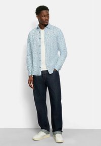 Hellblaues, gemustertes Hemd zum zuknöpfen, kombiniert mit einem weißen T-Shirt und dunklen Jeans. Abgerundet mit weißen Turnschuhen und hochgekrempelten Ärmeln.