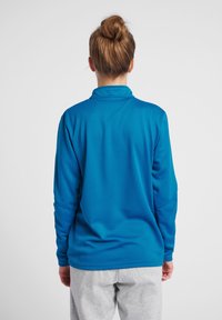 Maglione a maniche lunghe in un tessuto blu brillante con una superficie texture e un alto colletto, abbinato a pantaloni della tuta grigi chiari.