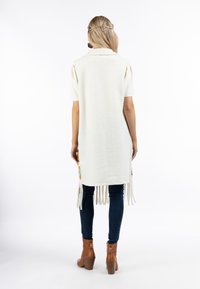 Langer weißer Weste mit fransigem Saum, strukturiertem Stoff, kombiniert mit dunklen Jeans und braunen Stiefeletten. Gestylt mit einem einfachen Kragen und ohne Muster.