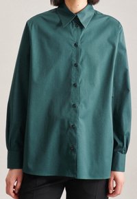 Turkos skjorta med knappar, spetsig krage, långa ärmar och svarta knappar. Tyget har en slät struktur och en avslappnad passform.