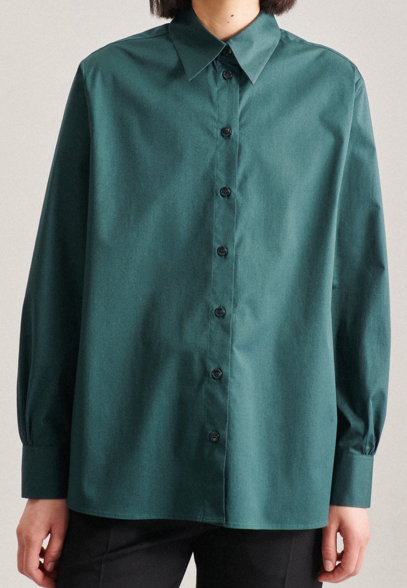 Turkos skjorta med knappar, spetsig krage, långa ärmar och svarta knappar. Tyget har en slät struktur och en avslappnad passform.