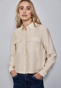 Camicia button-up beige con colletto e finitura testurizzata, dotata di due tasche frontali e maniche lunghe. Abbinata a jeans blu scuro.