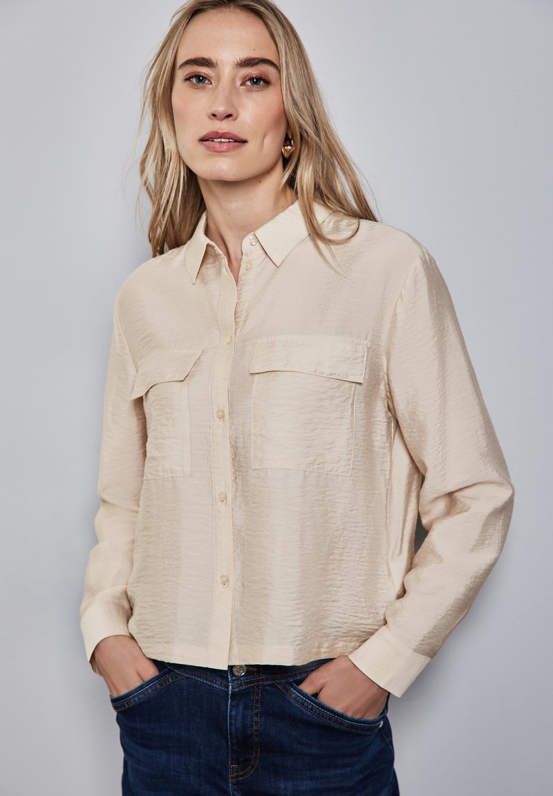 Camicia button-up beige con colletto e finitura testurizzata, dotata di due tasche frontali e maniche lunghe. Abbinata a jeans blu scuro.